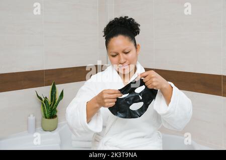 Charmante Frau in weißem Bademantel Anwendung feuchtigkeitsspendende schwarze Bettmaske während der Hautpflege Routine am Morgen Stockfoto