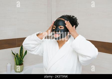 Charmante Frau in weißem Bademantel Anwendung feuchtigkeitsspendende schwarze Bettmaske während der Hautpflege Routine am Morgen Stockfoto