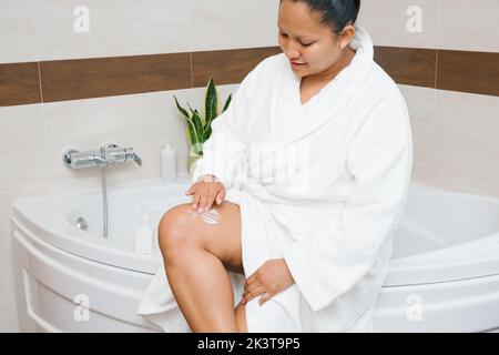 Fröhliche hispanische Frau, die feuchtigkeitsspendende Körpercreme auf die Beine anwendet, während sie im Badezimmer eine Hautpflege macht Stockfoto
