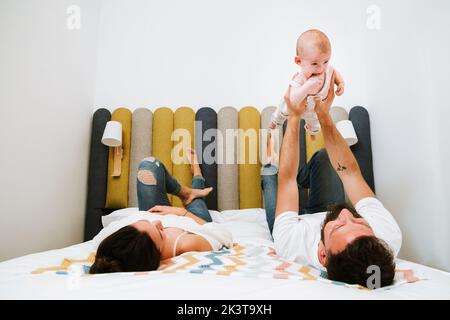 Vater gibt vor, dass verspielte Baby fliegen wie Superheld, während zusammen mit Frau und Kind auf weichem Bett zu Hause chillen Stockfoto