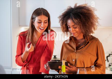 Positive junge multirassische Frauen Freunde in hellen Outfit Browsing Tablet während zusammen in modernen hellen Küche stehen Stockfoto