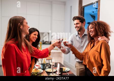 Seitenansicht von fröhlichen, vielfältigen Freunden, die feiern und mit Weinläsern klatschen, während sie am servierten Tisch mit leckerem Abendessen zu Hause stehen Stockfoto
