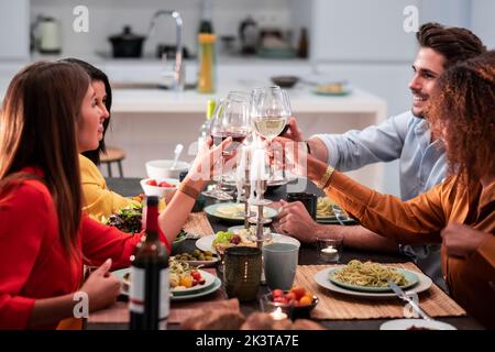 Seitenansicht von fröhlichen, vielfältigen Freunden, die feiern und mit Weinläsern klatschen, während sie am servierten Tisch sitzen und zu Hause ein köstliches Abendessen genießen Stockfoto