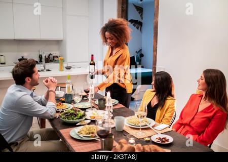 Schwarze afroamerikanische Frau serviert Wein zu fröhlichen Jugendlichen verschiedenen Freunden lachen und genießen Zeitvertreib, während sich beim Abendessen in hom Stockfoto