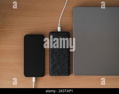 Ein Telefon, das von einer Powerbank neben einem Laptop auf einem Holztisch aufgeladen wird. Stockfoto