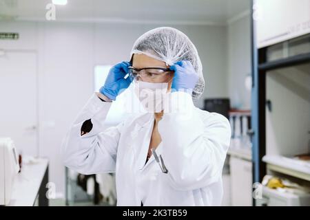 Seitenansicht der Frau in Uniform, die eine Brille auf das Gesicht legt, während sie sich auf die Operation im Labor vorbereitete Stockfoto