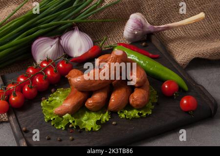 Leckere Räucherwürste auf einem Holzbrett, mit frischem Gemüse dekoriert Stockfoto