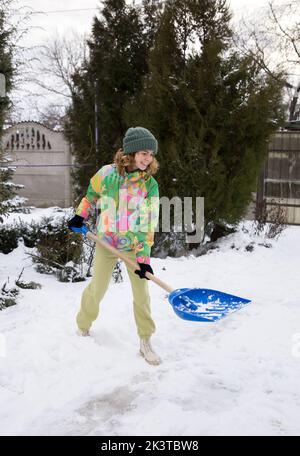 Positives Teenager-Mädchen in strahlend warmer Sportkleidung, räumt nach einem starken Schneesturm den Bürgersteig, die Straße, den Hof vom Schnee ab, wirft den Schnee mit einem großen Sh ab Stockfoto