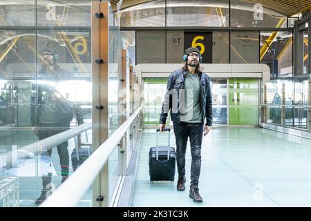 Hipster-Mann in schwarzer Lederjacke und Jeans mit Kopfhörern, während er mit dem Koffer in der Flughafenhalle läuft Stockfoto