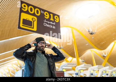 Hübscher bärtiger Hipster mit Brille und Kopfhörer in schwarzer Lederjacke und Strickmütze, der in der Flughafenhalle steht und wegschaut Stockfoto