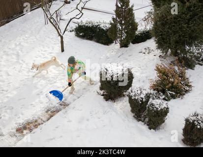 Junge Frau in strahlend warmer Sportkleidung, die nach einem starken Schneesturm Bürgersteig, Straße, Hof vom Schnee räumt, wirft Schnee auf Schaufel zur Seite. Wintersaison Stockfoto