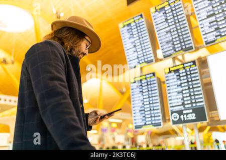 Von der unteren Seite sehen Sie einen erwachsenen bärtigen männlichen Reisenden in stilvollem Outfit, der sein Mobiltelefon hält und Informationen über den Flug im Flugplan eincheckt Stockfoto