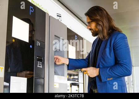 Seitenansicht eines eleganten Geschäftsmannes mit Koffer, der am Parkplatzsterminal an der Bezahlstation sitzt und am Flughafen bezahlt Stockfoto