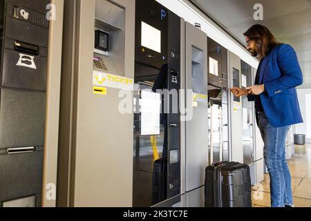 Von unten sehen Sie einen stilvollen Geschäftsmann mit Koffer, der den Parkplatzparkplatz am Bezahlterminal benutzt und am Flughafen bezahlt Stockfoto