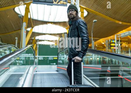 Von unten sehen Sie den trendigen bärtigen erwachsenen Mann in Hut und Lederjacke mit Koffer, der auf der Rolltreppe im Flughafen steht Stockfoto