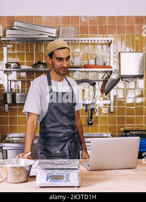 Positiver junger ethnischer männlicher Bäckereibesitzer, der an der Theke mit Laptop und Zutaten für die Backarbeit steht Stockfoto