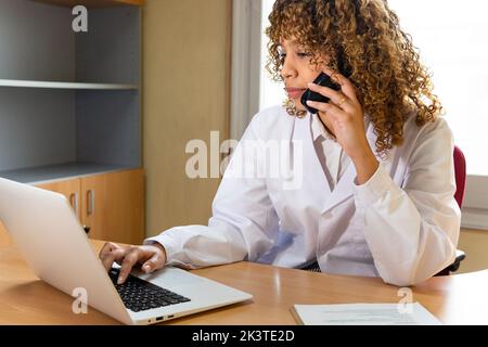Crop ethnische weibliche Sanitäter arbeiten auf tragbaren Computer, während auf Telefon Hörer am Tisch im Büro sprechen Stockfoto