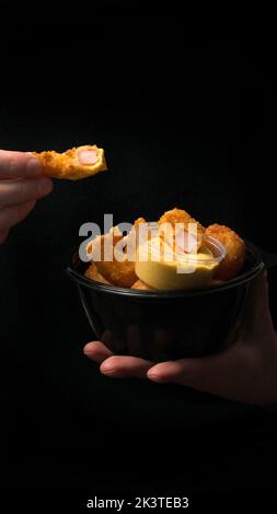 Ein junger Mann isst leckere panierte Garnelen mit Käsesauce auf schwarzem Hintergrund Stockfoto