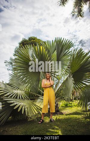 In voller Länge Inhalt junge Frau in gelbem Jumpsuit, die Kamera anschaut und Haare berührt, während sie in der Nähe exotischer grüner Pflanzen im Sommerresort steht Stockfoto