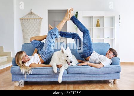 Seitenansicht des positiven Freundes und der Freundin, die auf dem Sofa liegen und die Füße berühren, während sie sich am Wochenende mit dem weißen Schweizer Schäferhund im Zimmer amüsieren Stockfoto