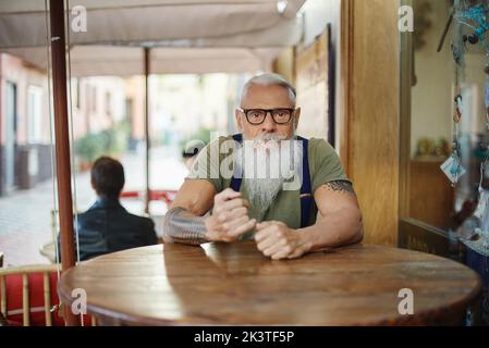 Älterer, stilvoller bärtiger Dandy-Mann mit Tätowierung und Brille, der am Cafeteria-Tisch auf die Kamera schaut Stockfoto