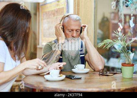 Crop lächelnde Enkelin Textnachrichten auf Handy in der Nähe fröhlich stilvollen Dandy Opa Musik aus Headset am Café-Tisch hören Stockfoto