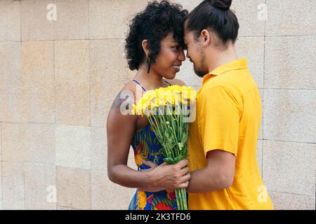 Liebevoller ethnischer Mann umarmt lächelnde Frau mit Blumenstrauß, während er im Sommer auf der Straße steht Stockfoto