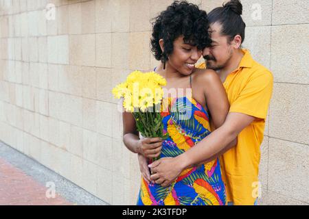 Liebevoller ethnischer Mann umarmt lächelnde Frau mit Blumenstrauß, während er im Sommer auf der Straße steht Stockfoto