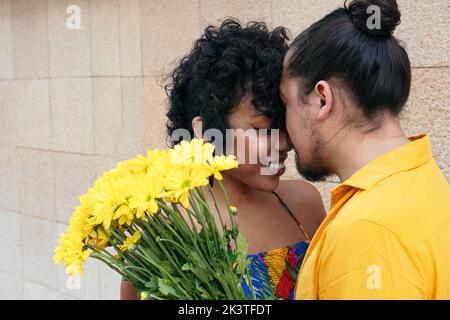 Liebevoller ethnischer Mann umarmt lächelnde Frau mit Blumenstrauß, während er im Sommer auf der Straße steht Stockfoto