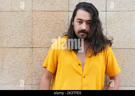 Bärtiger Hipster mit langen, welligen Haaren und gelbem Sommerhemd, der an der Wand auf der Straße steht und wegschaut Stockfoto