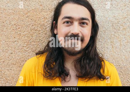 Bärtiger Hipster mit langen, welligen Haaren und gelbem Sommerhemd, der auf der Straße gegen die Wand steht und die Kamera anschaut Stockfoto