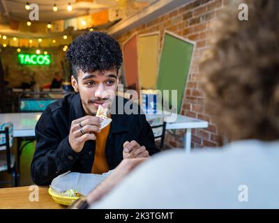 Lächelnder ethnischer Mann mit köstlichem Taco, der am Restauranttisch mit einem unverkennbaren männlichen Partner interagiert Stockfoto