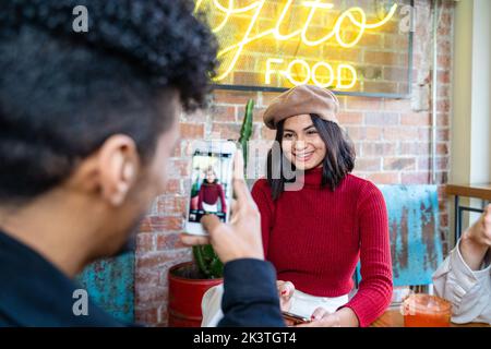Mann mit Smartphone, der in einem modernen Restaurant ein Bild von einer fröhlichen, lächelnden ethnischen Freundin macht Stockfoto