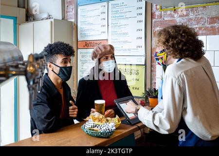 Junge ethnische Paare in Masken, die Essen bestellten und mit männlichen Kellner sprachen, die im modernen Restaurant an der Theke standen Stockfoto
