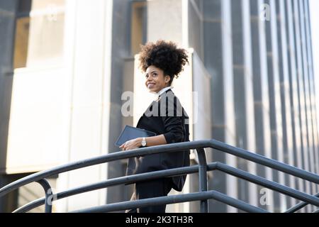 Seitenansicht aus dem niedrigen Blickwinkel einer lächelnden afroamerikanischen Architektin im Anzug, die mit einem Tablet in der Stadt steht und wegschaut Stockfoto
