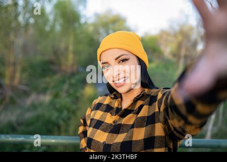 Inhalt junge lateinamerikanische Touristin in kariertem Hemd und Hut lächelnd und schauend auf die Kamera, während sie auf einem hölzernen Steg über einem schmalen R steht Stockfoto