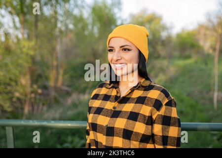 Inhalt junge lateinamerikanische Touristin in kariertem Hemd und Hut lächelnd und wegblickend, während sie auf einem hölzernen Steg über dem schmalen Fluss steht Stockfoto
