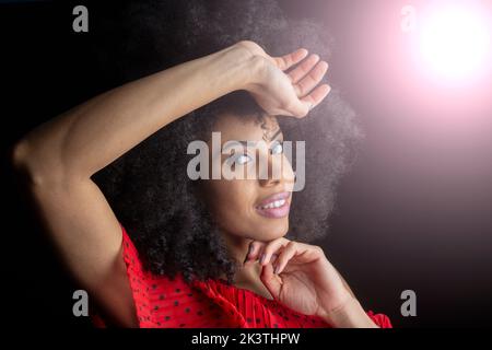 Seitenansicht einer fröhlichen jungen ethnischen Frau mit Afro-Haaren in ornamentaler roter Kleidung mit Make-up und Afro-Frisur, die in schwarzer Studio-BA auf die Kamera schaut Stockfoto