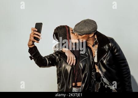 Afroamerikanischer Mann und Frau in stylischen Outfits küssen sich gegenseitig und machen Selfie, während sie an der grauen Wand auf der Straße stehen Stockfoto