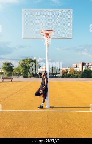 Müde afroamerikanische Sportler in Uniform lehnt neben dem Korb auf gelb markierten Spielplatz mit Ball in hellen Tag Stockfoto