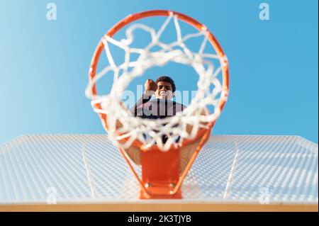 Von unten sehen Sie den afroamerikanischen Sportler, der auf einem Basketballring steht und mit einer geballten Faust im Siegessymbol auf die Kamera blickt Stockfoto