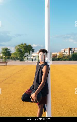 Müde afroamerikanische Sportler in Uniform lehnt neben dem Korb auf gelb markierten Spielplatz mit Ball in hellen Tag Stockfoto