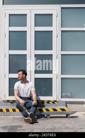 Mann mit künstlichem Bein sitzt auf Schritt in der Nähe von Skateboard an sonnigen Tag auf der City Street Stockfoto