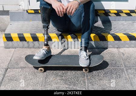 Von oben anonymer Mann mit künstlichem Bein sitzt auf Schritt in der Nähe von Skateboard an sonnigen Tag auf der Straße der Stadt Stockfoto