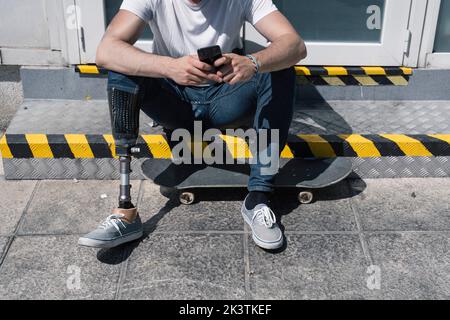 Von oben anonymer Mann mit künstlichem Bein Browsing Smartphone während auf Schritt in der Nähe von Skateboard an sonnigen Tag auf der Straße der Stadt sitzen Stockfoto