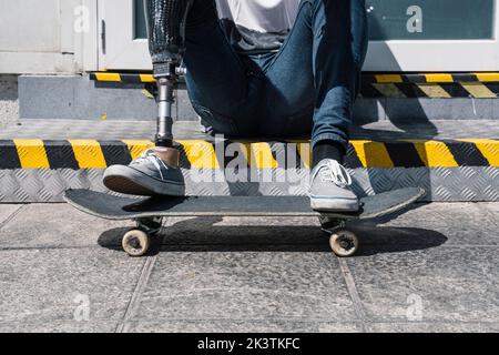 Von oben anonymer Mann mit künstlichem Bein sitzt auf Schritt in der Nähe von Skateboard an sonnigen Tag auf der Straße der Stadt Stockfoto