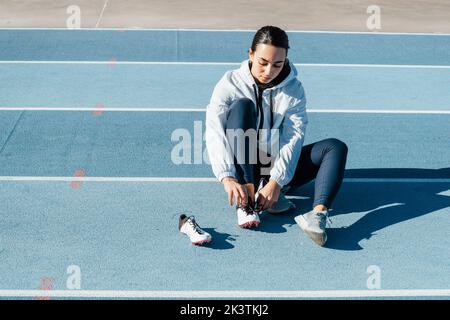 Sportlerin in Sportbekleidung sitzt auf der Rennstrecke und wechselt die Schuhe während des Trainings auf der Rennstrecke an einem sonnigen Tag im Stadion Stockfoto