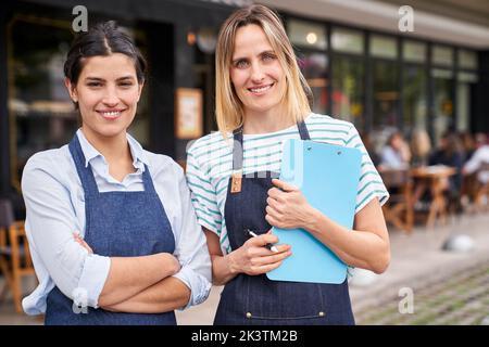 Mittellange Aufnahme von zwei weiblichen Restaurantbesitzern, die vor ihrem Restaurant stehen und die Kamera betrachten Stockfoto