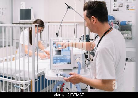 Seitenansicht des Arztes männliche Einstellparameter medizinisches Gerät für die künstliche Atmung in der Station mit Krankenschwester untersucht Baby auf dem Hintergrund Stockfoto