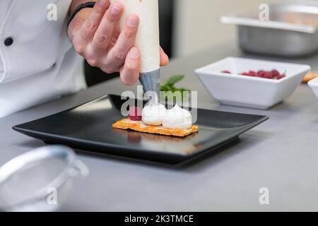 Crop man in weißer Chefjacke schmückt ausgefallene Gerichte mit Zuckerguss aus Paspelsack und Himbeere auf dem Teller in der Restaurantküche Stockfoto